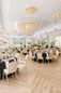 Elegancka sala weselna z jasną aranżacją stołów Przestronna sala weselna z okrągłymi stołami, jasnymi krzesłami i eleganckimi dekoracjami kwiatowymi.